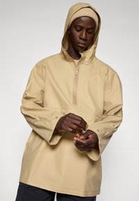 Helle beige wasserdichte Pulloverjacke mit halber Reißverschlusskragen, verstellbaren Bündchen und Kapuze. Glatte Textur, lockere Passform.