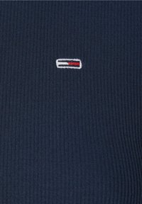 Marinblå ribbad tyg med en liten broderad logotyp med röda och vita accenter, centrerad i designen.