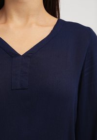 Marineblaue Bluse mit V-Ausschnitt und subtiler Textur. Verfügt über ein tonales Knopfdetail am Hals und langen Ärmeln. Leichte Stoffqualität.