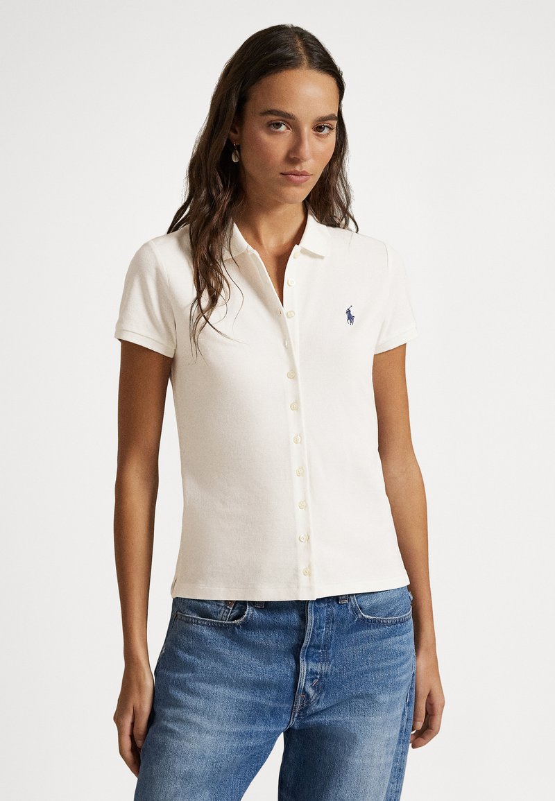 Polo branco de manga curta com frente abotoada e pequeno logótipo azul no peito esquerdo, combinado com jeans azuis. Tecido de textura suave.