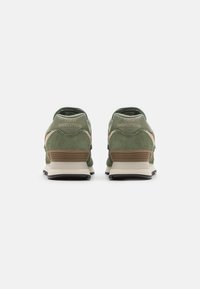 Zielone zamszowe buty biegowe z beżowym akcentem, mające czarne podeszwy i teksturowany wzór na pięcie. Logo "New Balance" widoczne z tyłu.