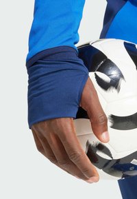 Blaues, langärmliges Sportshirt mit navyfarbigen Manschetten, gehalten von einer Hand, die einen schwarz-weißen Fußball mit markanten Mustern umschließt.