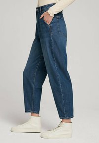 Jeans z wysokim stanem w niebieskim denimie, o luźnym kroju, z przednimi kieszeniami i zwężanymi nogawkami. Noszone z białymi trampkami za kostkę.