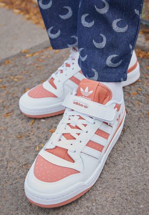 Baskets Adidas blanches et corail avec bride Velcro portées avec un pantalon bleu à motifs sur un trottoir jonché de feuilles mortes.
