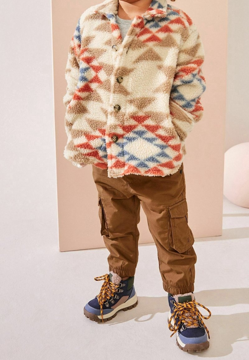 Enfant portant une veste polaire boutonnée aux motifs rouges, bleus et beiges, un pantalon cargo marron, et des chaussures de randonnée bleues avec des lacets jaunes.