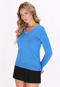 Blauer Pullover mit langen Ärmeln und V-Ausschnitt, mit einem weißen Besatz entlang des Ausschnitts und einem gerippten Saum. Tragen mit einem schwarzen, plissierten Rock.