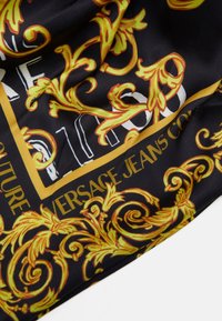 Černý šál s vzorem obsahující zlaté a žluté zdobené motivy s bílým nápisem "Versace Jeans Couture" a dalšími dekorativními prvky.