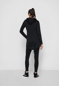 Donna con coda di cavallo che indossa una lunga felpa nera con cappuccio, leggins neri e sneakers alte nere, in piedi con le spalle alla macchina fotografica.