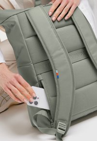 Zaino verde imbottito con strap regolabile e tasca esterna. Materiale liscio e resistente; piccolo dettaglio colorato vicino allo strap.