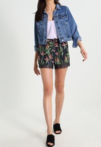 Žena v modré džínové bundě, bílé košili, květovaných šortkách s krajkovým lemem a černých otevřených pantoflích jde na bílém pozadí.