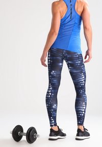 Débardeur de sport bleu et leggings noirs à motifs, présentant un dégradé avec des textures subtiles, accompagnés de chaussures d'entraînement noires et d'un haltère.