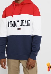 Felpa con cappuccio a blocchi di colore rosso, bianco e blu navy realizzata in tessuto morbido. Presenta una tasca a marsupio, un cappuccio regolabile con coulisse e il logo "TOMMY JEANS".