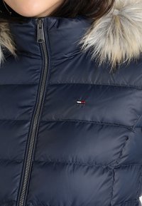 Námořnicky modrá péřová bunda s lesklým povrchem, vybavená odnímatelným šedým umělým kožešinovým límcem a zipem na zapínání, zdobená malým logem na hrudi.