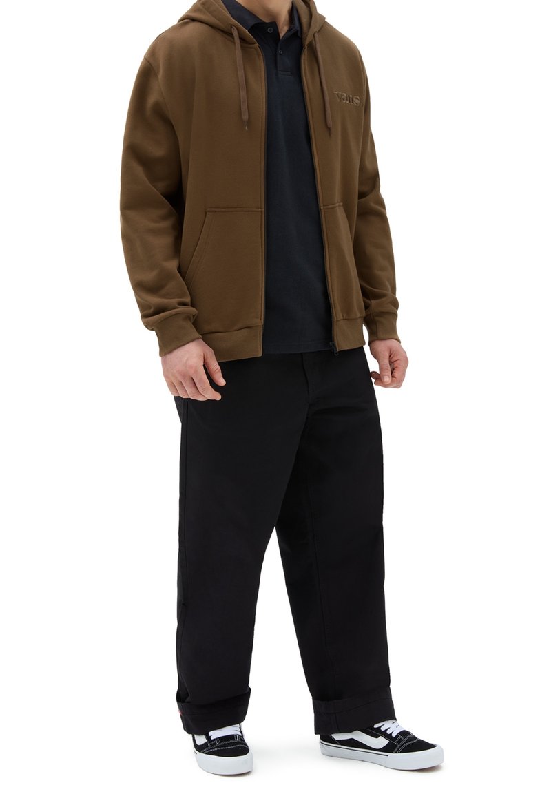 Homme portant un sweat à capuche marron zippé par-dessus un polo noir, un pantalon large noir et des baskets noir et blanc, debout sur fond blanc.