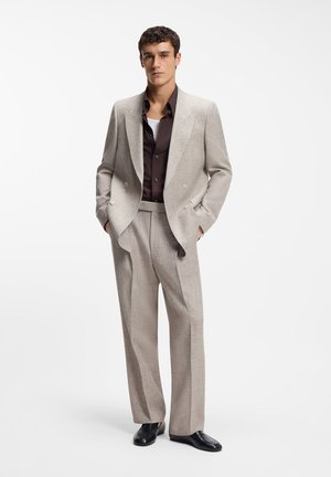 Mies, joka pukeutunut beigeen kaksiriviseen pukuun, tummanruskea paita, valkoinen aluspaita ja mustat kengät, seisoo kädet taskuissa.