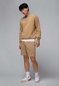 Beige mikina a odpovídající šortky vyrobené z měkkého materiálu, zdobené bílým logem. Model má na sobě bílé tenisky a vrstvené ponožky.
