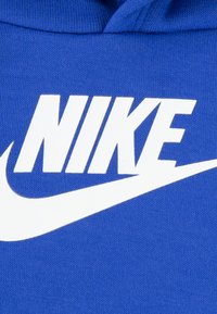 Zils džemperis ar baltu "NIKE" logotipu un swoosh dizainu. Audums ir gluds ar adījuma struktūru. Ir regulējama kapuce.