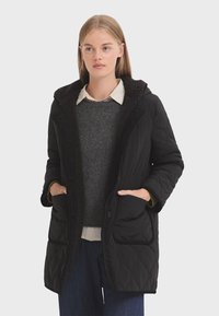 Manteau matelassé noir avec une capuche en fourrure marron texturée, deux poches avant et une fermeture à boutons. Porté sur un pull gris et un jean.