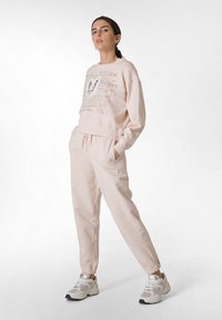 Jasnoróżowy sweatshirt z graficznym napisem, w komplecie z pasującymi dresami. Dresy mają elastyczne mankiety i sznurek w pasie. Uzupełnione białymi sneakersami.