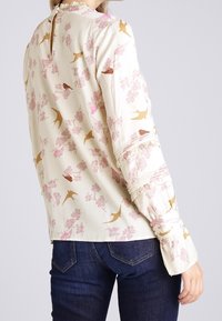 Krämfärgad blus med fågel- och blommönster. Har långa ärmar, gathered axlar och en nyckelhålrygg. Strukturerad tyg, mjukt avslut.