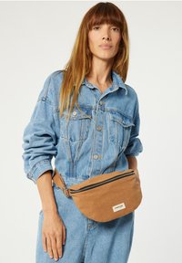 Banane en tissu marron avec une forme arrondie, fermeture éclair sur le dessus et écusson avec logo ; portée par-dessus une tenue en denim à manches longues.