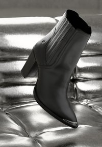 Černé kožené kotníkové boty s elegantním designem, špičatou špičkou, vysokým silným podpatkem a vertikálními žebrovanými detaily na stranách. Lesklý povrch.