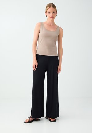 Woman standing wearing a beige sleeveless top, black wide-leg pants, black sandals, and gold round earrings on a plain light background.