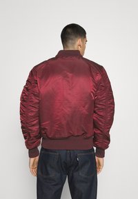 Giacca bomber in satin bordeaux con polsini e orlo a costine, dotata di maniche gatherate e una texture liscia e lucida. Vista posteriore mostrata.