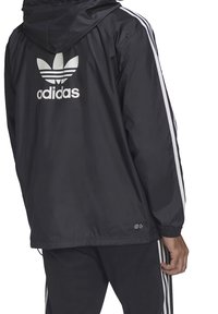 Černá větrovka s kapucí, s bílým logem Adidas a třemi bílými pruhy podél rukávů. Lehká, hladká textura.