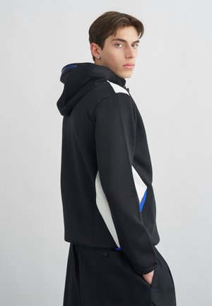 Noor mees, kes on näoga küljele, kannab musta kapuutsiga pusa valgete ja siniste detailidega, käed taskutes heleda ühevärvilise taustaga.