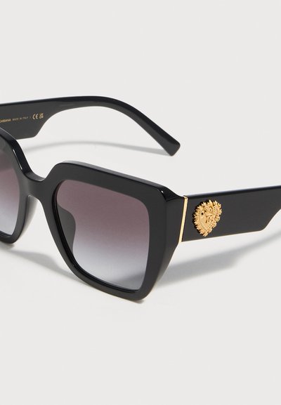 Μαύρα τετράγωνα γυαλιά ηλίου μεγάλου μεγέθους με φακούς με διαβάθμιση και χρυσό διακοσμητικό έμβλημα στο πλαινό μέρος, τοποθετημένα σε λευκό φόντο.