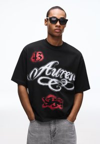 Fekete pamut póló nagy fehér és piros grafikával, amelyen a 43-as szám és egy piros oroszlánembléma látható, szürke farmer rövidnadrággal viselve.