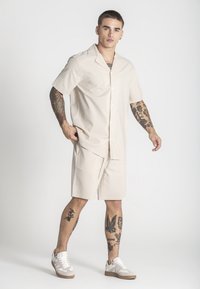Camisa de botones de manga corta beige y pantalones cortos a juego, fabricados en tejido ligero. Presenta un corte relajado, con bolsillos laterales visibles.