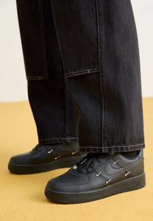 Czarne skórzane sneakersy z detalami z srebrnych agrafek noszone z luźnymi czarnymi spodniami na beżowej podłodze.