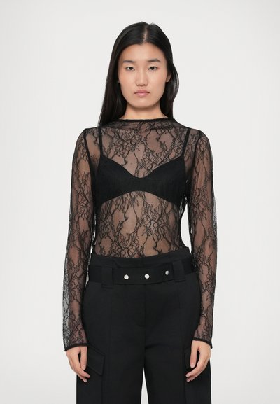 Donna che indossa un top in pizzo nero a maniche lunghe sopra un reggiseno nero, abbinato a pantaloni neri a vita alta con una cintura larga e bottoni argentati.