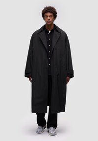 Abrigo largo negro hecho de una tela suave y ligera, con un amplio collar, bolsillos laterales y un corte relajado. Llevado sobre una camisa y pantalones negros.