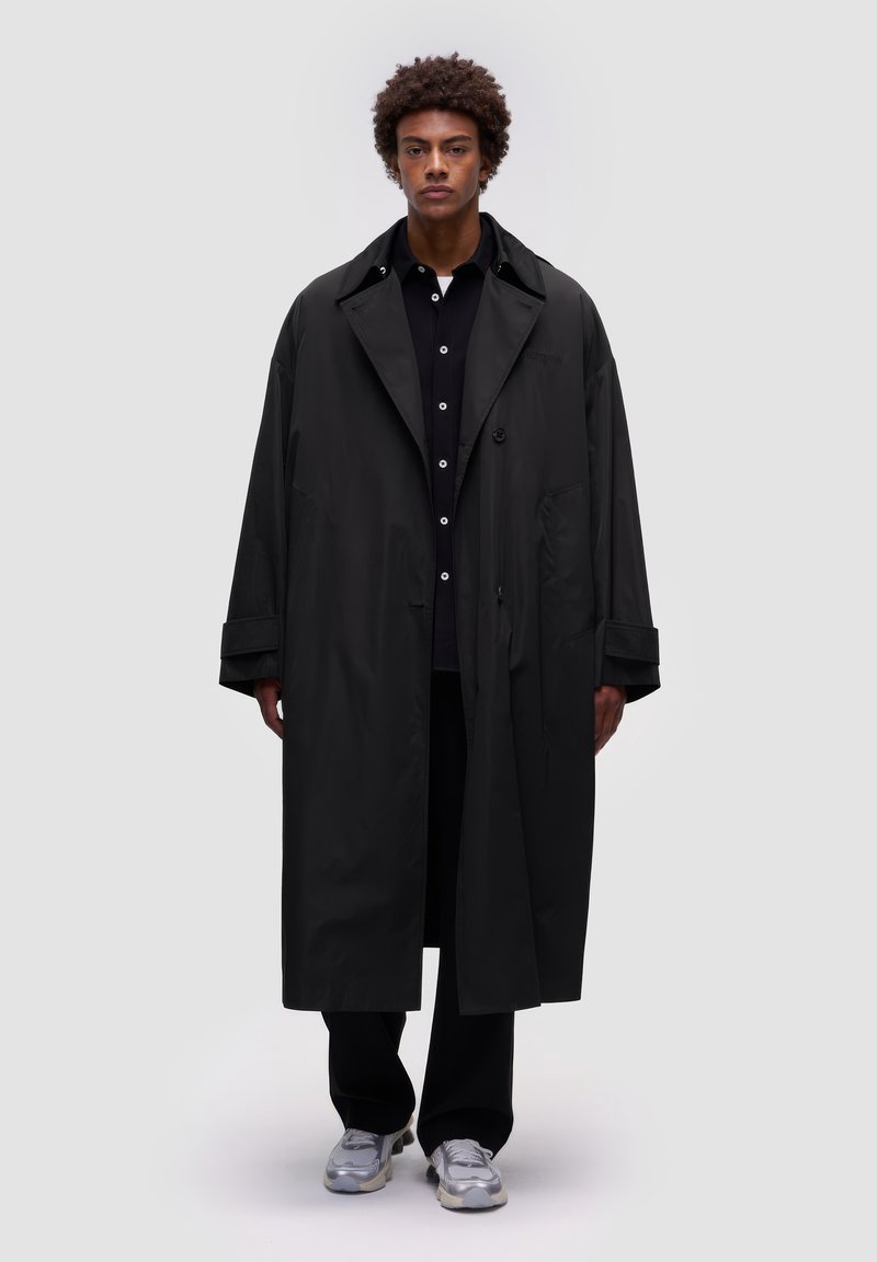 Abrigo largo negro hecho de una tela suave y ligera, con un amplio collar, bolsillos laterales y un corte relajado. Llevado sobre una camisa y pantalones negros.