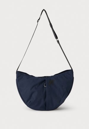Sac à bandoulière bleu marine en forme de demi-lune avec sangle noire réglable et logo noir discret sur le panneau avant sur fond blanc.