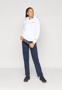 Λευκό φούτερ με κουκούλα και μπροστινή τσέπη με κεντημένο λογότυπο, συνδυασμένο με μπλε σκούρο φόρμας παντελόνι και μαύρα αθλητικά παπούτσια, κρατώντας ένα μπουκάλι νερό.