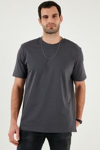 Muškarac obučene tamnosive kratke majice, crnih traperica, srebrne ogrlice i crnih narukvica, stoji ispred čistog bijelog zida.