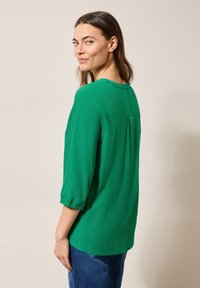 Blusa verde con mangas tres cuartos, tela suave, escote redondo y un pliegue en la parte posterior. Usada con pantalones de mezclilla azul.