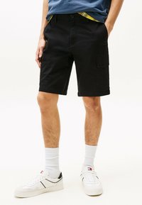Pantalones cortos cargo negros con frente plano, dos bolsillos laterales y longitud hasta la rodilla. Llevados con zapatillas blancas y calcetines blancos acanalados.