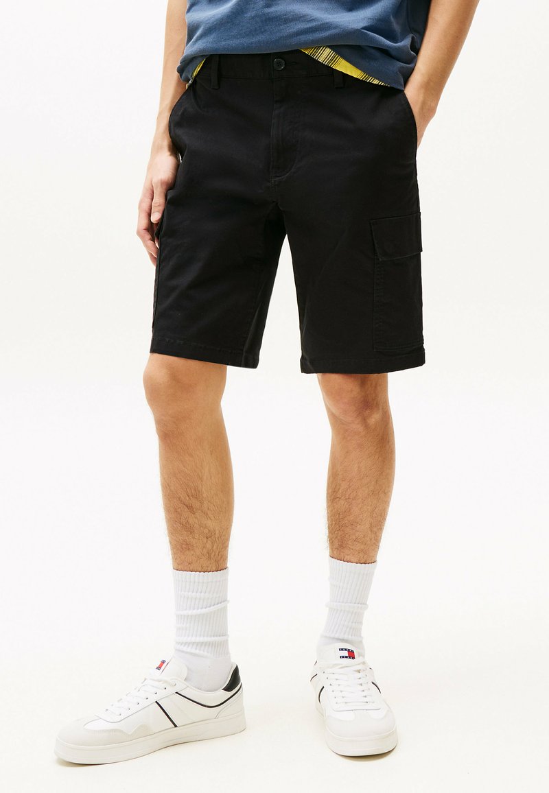 Pantalones cortos cargo negros con frente plano, dos bolsillos laterales y longitud hasta la rodilla. Llevados con zapatillas blancas y calcetines blancos acanalados.