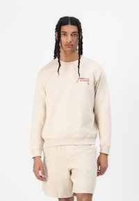 Beige sweatshirt with ribbed cuffs and hem, featuring red printed text on the chest. Paired with matching beige shorts.