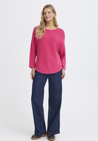 Rózsaszín kötött pulóver laza szabással, kerek nyakkivágással és hosszú ujjakkal, széles szárú sötétkék farmernadrággal és barna fűzős csizmával párosítva.