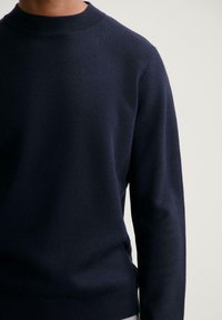 Hombre vistiendo un suéter de punto de manga larga en azul marino, con un cuello y dobladillo acanalados, contra un fondo claro y liso.