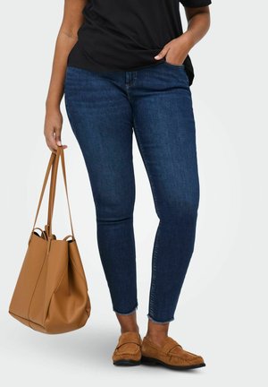 Osoba v tmavomodrých úzkych džínsoch, hnedých semišových mokasínach, čiernom topánke, nesúca v ľavej ruke veľkú hnedú koženú kabelu.