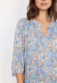 Blusa azzurra chiara con motivi floreali in colori pastello. Presenta un collo a V, una consistenza morbida e maniche a tre quarti.