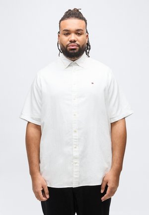 Homme aux cheveux tressés et barbe, portant une chemise blanche à manches courtes boutonnée et un pantalon noir, debout devant un fond blanc uni.
