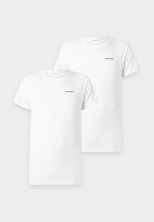 Mann steht barfuß, trägt ein weißes kurzärmeliges Hemd und schwarze Boxershorts vor einem schlichten weißen Hintergrund.